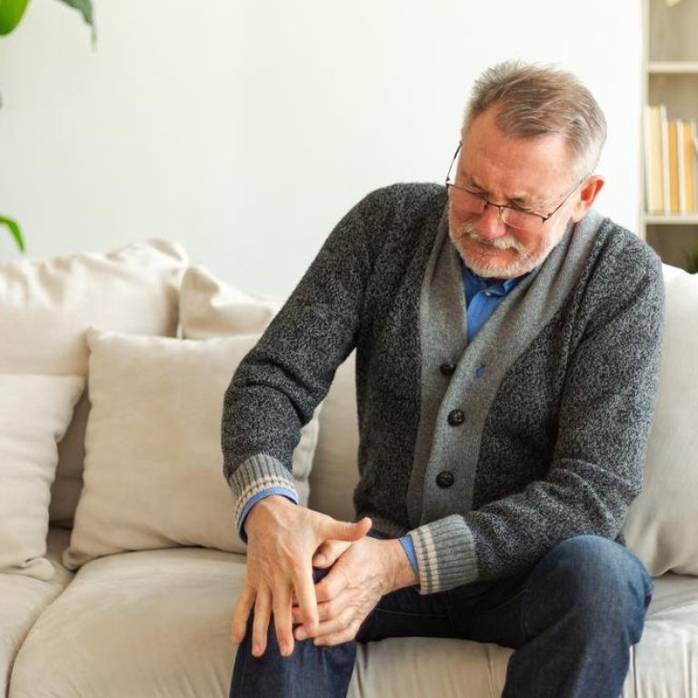 Knee pain arthritis body sick health care concept. Unhappy middle aged senior man suffering from knee ache sitting on sofa at home, Mature old senior grandfather touching leg feeling pain hurt in knee