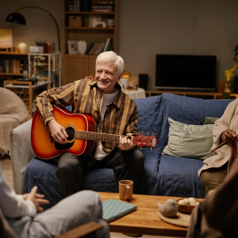 Group of seniors including Caucasian man playing acoustic guitar and smiling, senior Caucasian woman and senior Black woman sitting together and listening in nursing home common area
